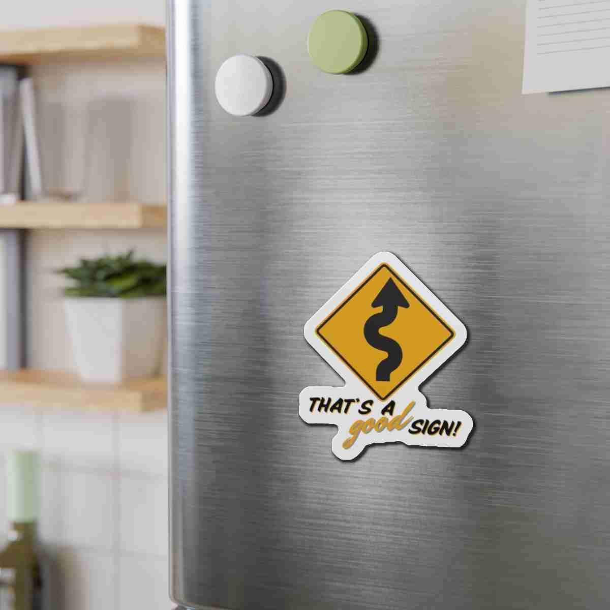 Stainless steel refrigerator door with a yellow winding-road sticker that reads 'That's a good sign!' in a kitchen.