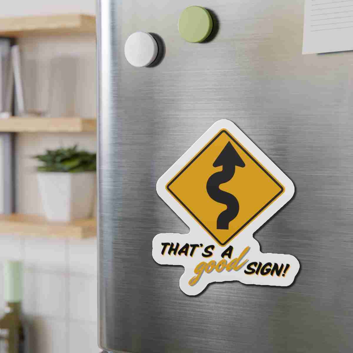 Stainless steel fridge door with a yellow diamond road-sign sticker featuring a winding road and the caption 'That’s a good sign!'