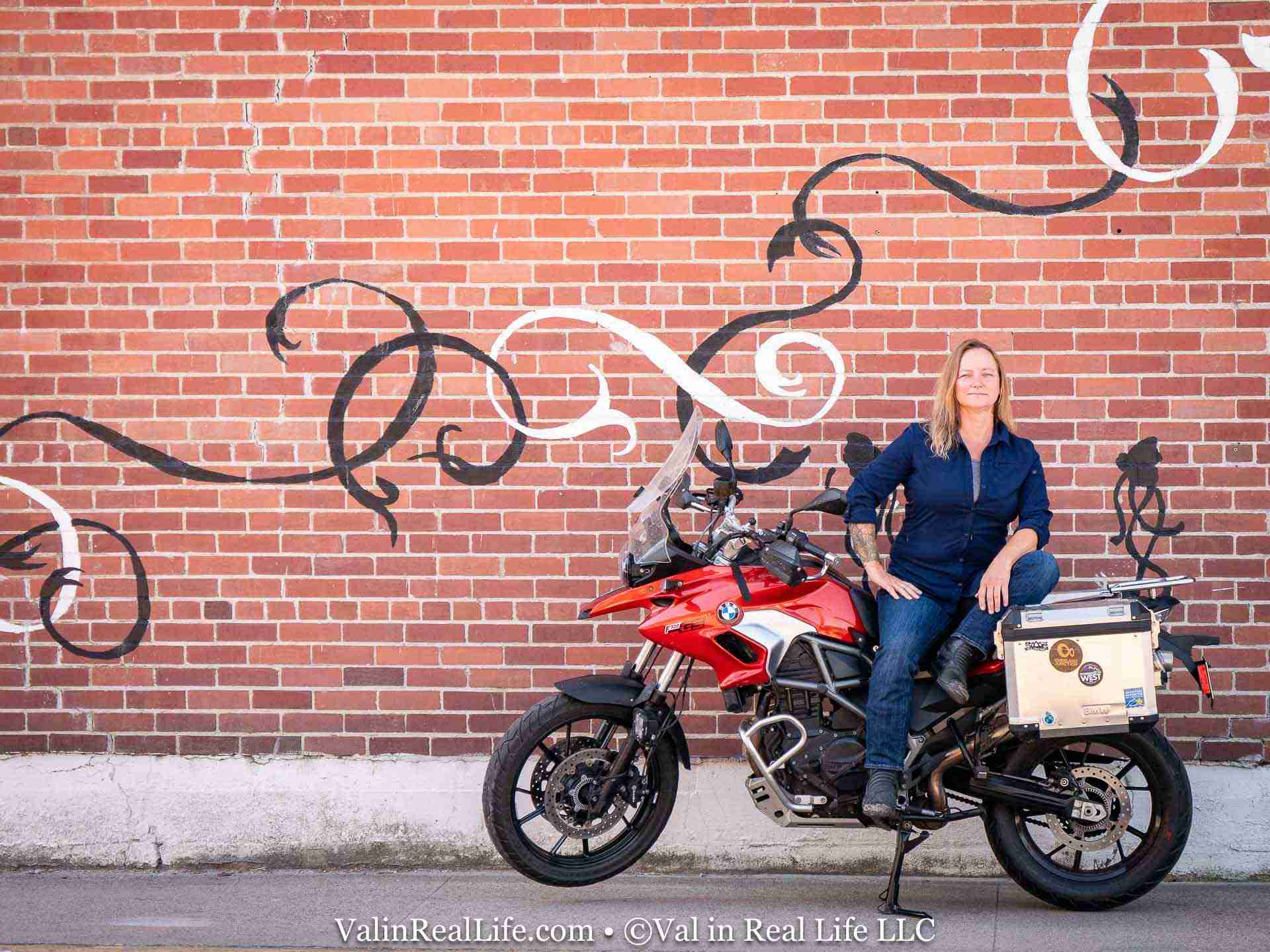 Woman sits on a red BMW adventure motorcycle in front of a graffiti-decorated brick wall outdoors.